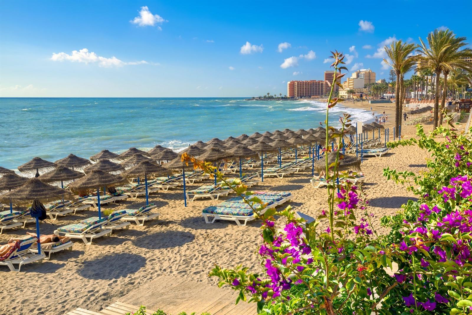 Straw umbrellas and loungers on Benalmádena beach, with bougainvillea in front and the hotels of the Costa behind