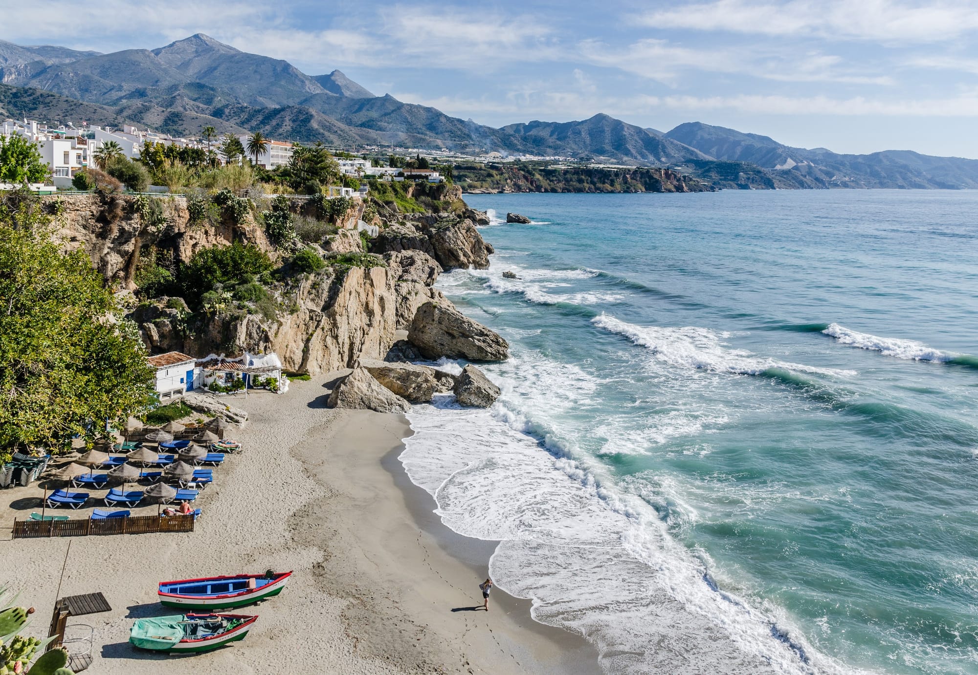 The view from the Balcón de Europa, Nerja — turquoise sea against wave-cut cliffs, the white town above, two fishing boats on an empty beach