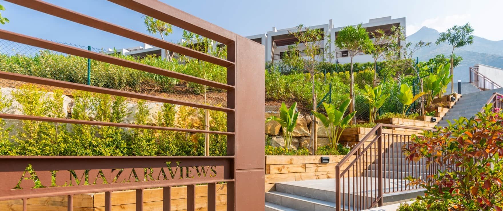 The entrance gate, with Almazara Views cut into its metal, and planted terraces and steps climbing to the houses. Almazara Views, Cerros del Lago, Istán.