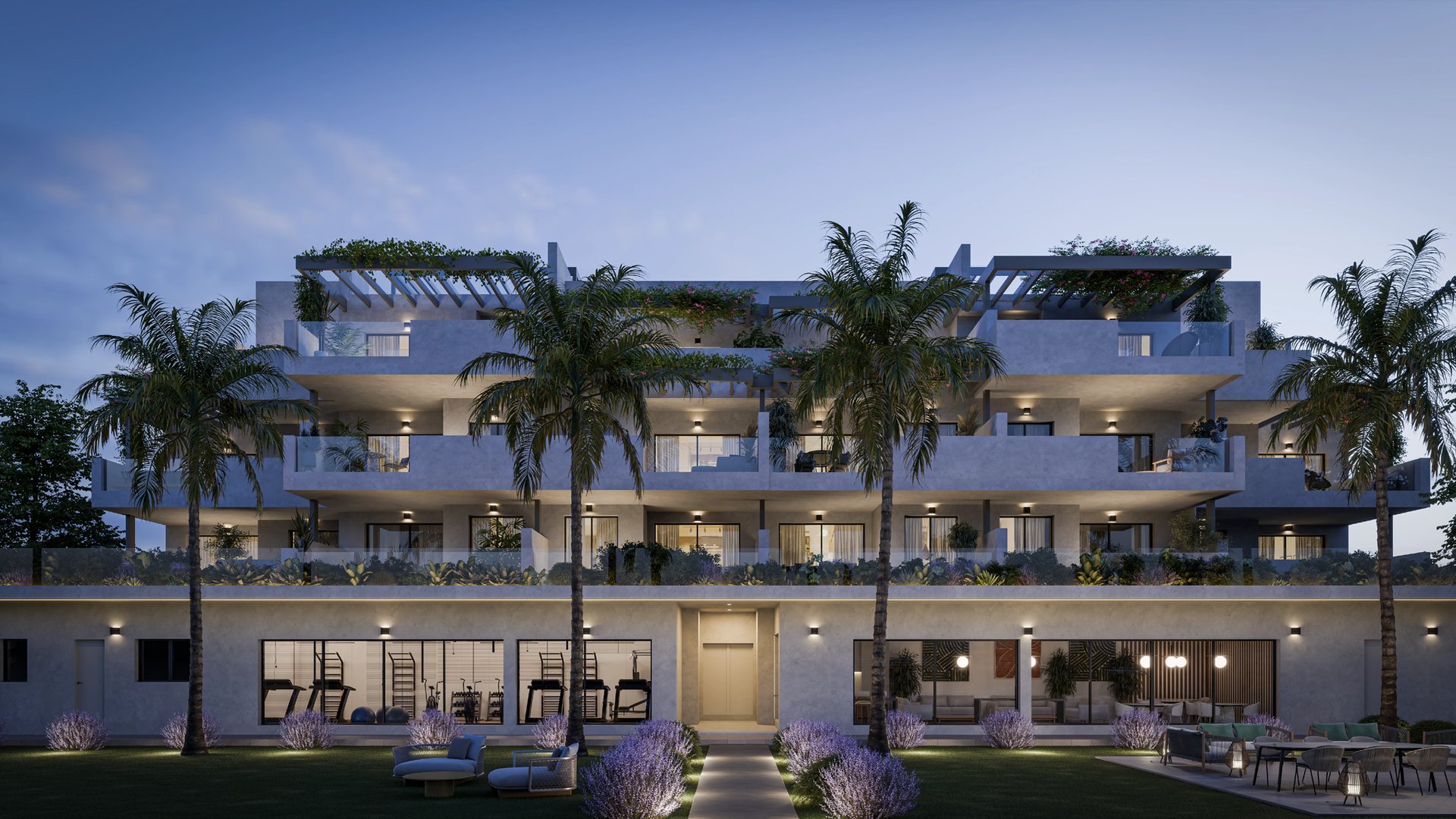 The front of one of the buildings at dusk, its terraces lit and stepped back above the ground floor gardens, with palms planted along the front. Alura Living, Casares Costa, Casares.