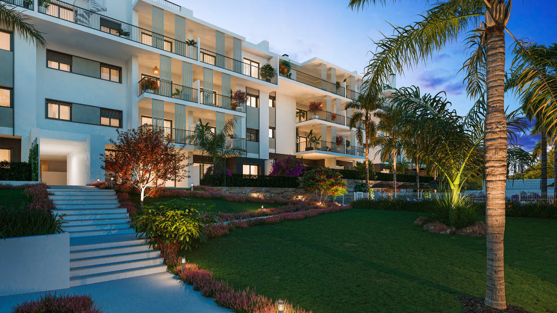 The garden between the buildings at dusk, a lawn and steps running between them and the windows lit above. Isidora Living, Calle Carlos Cano, Estepona.