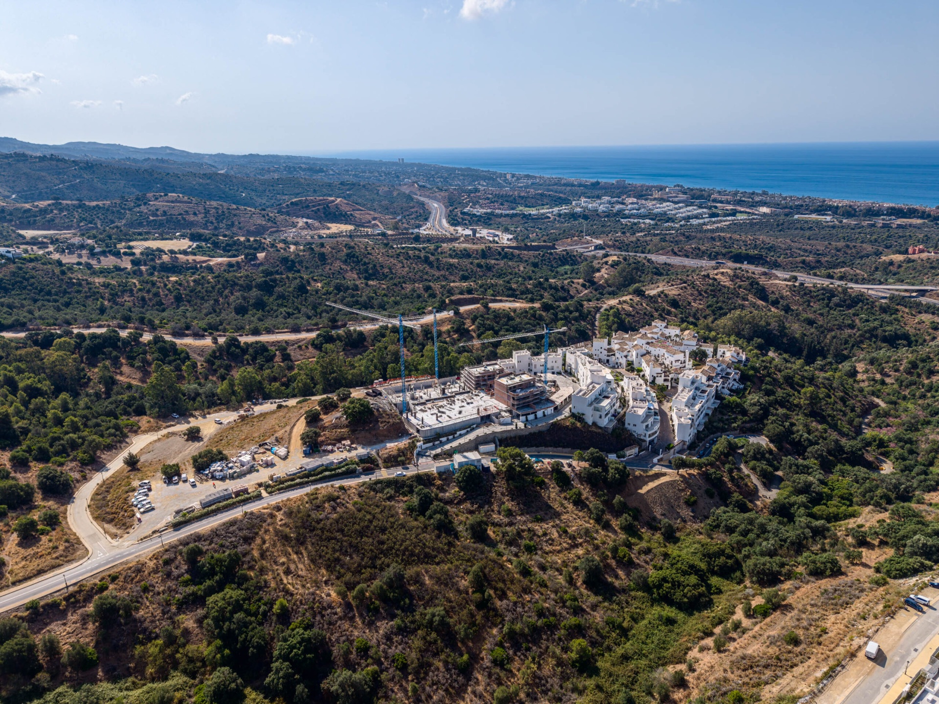 Two cranes still working over Origin in July 2026, with several of its buildings already finished in white and the coast beyond. Origin, Altos de los Monteros, Marbella.