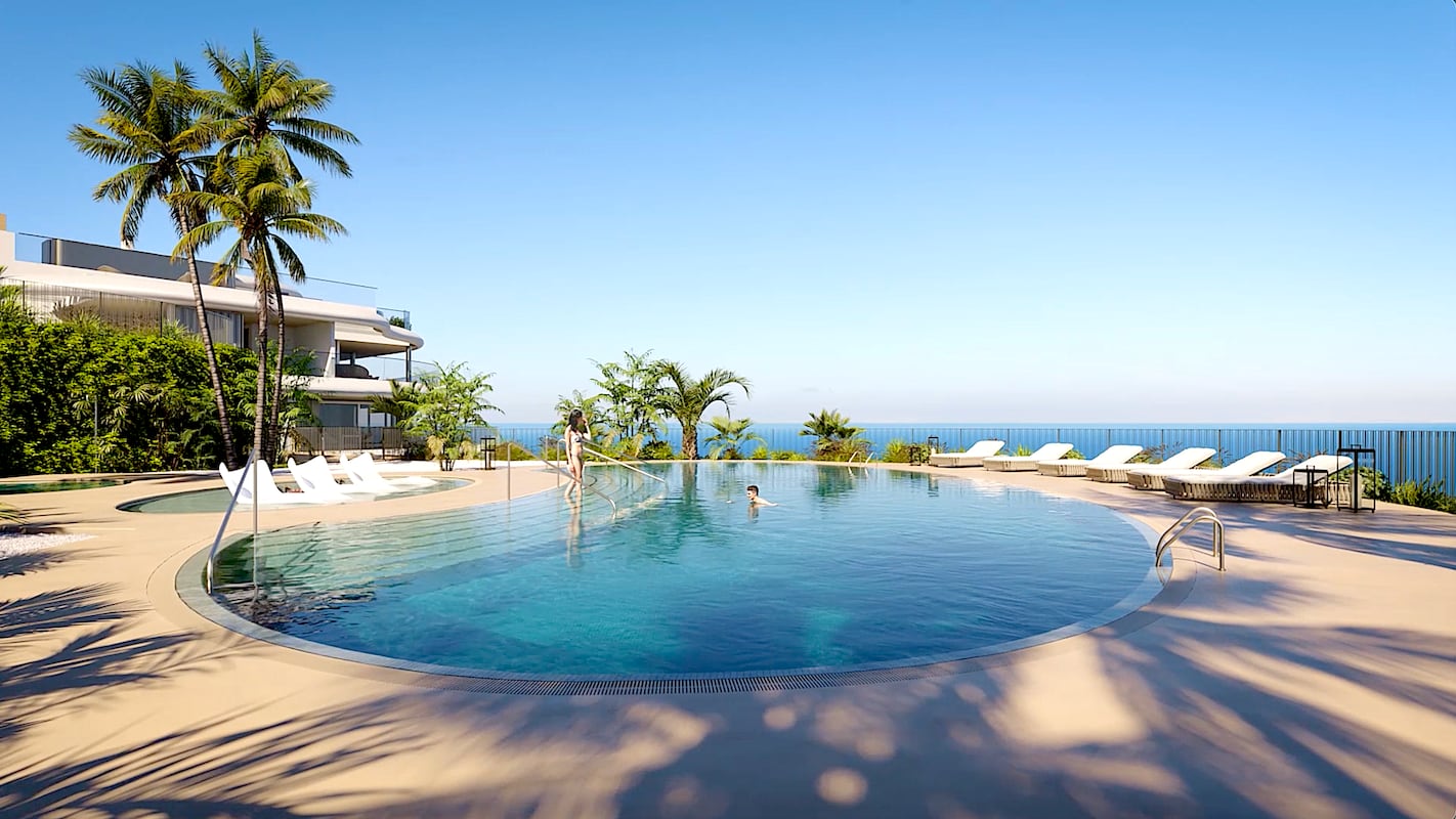 The shared pool, with loungers along its far side, an apartment building behind the palms on the left, and the sea on the horizon. The Sky Marbella, La Alborada, Benahavís.