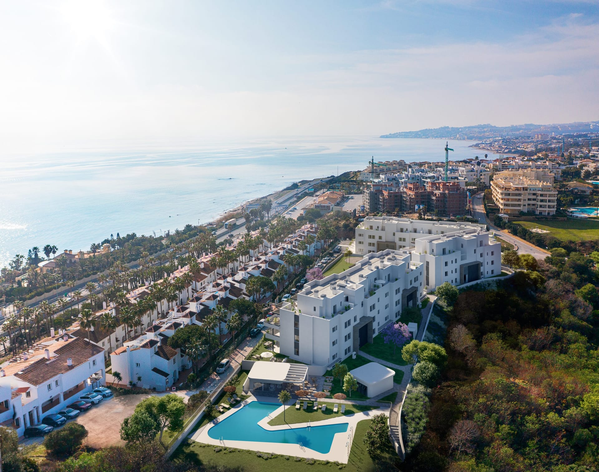 An aerial view over the pool and gardens to the buildings, with the coast stretching west beyond them. Vitta Marina, El Chaparral, Mijas.