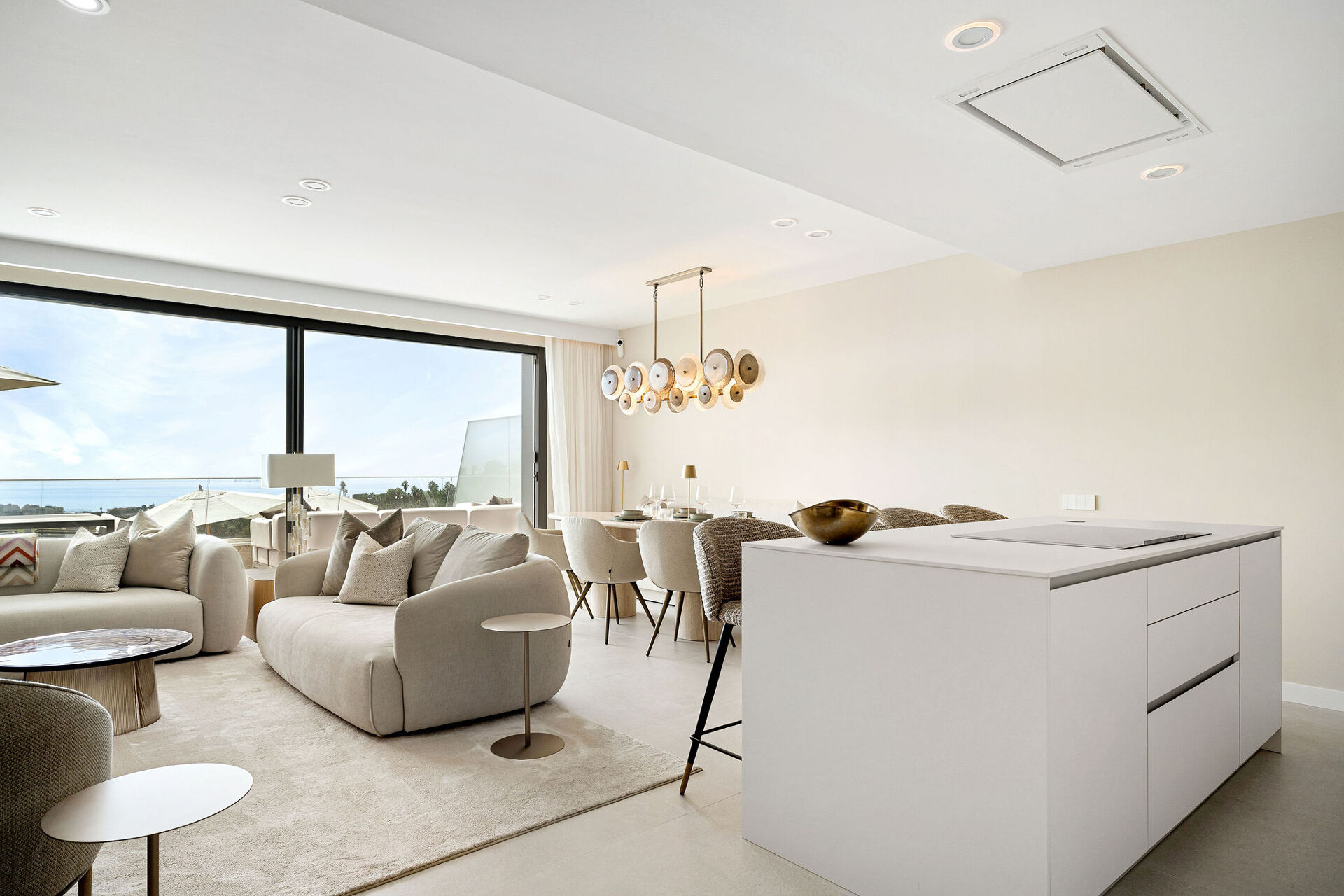 The open living space, with sofas by a wide window, a dining table under a pendant light and the kitchen island in front.