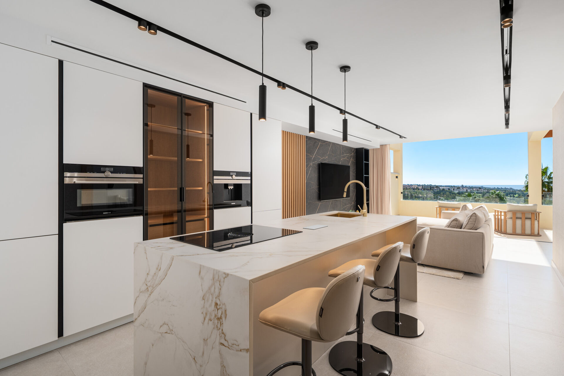 The kitchen island with a hob and a sink, Siemens ovens set into the tall white units, and the lounge and terrace beyond.