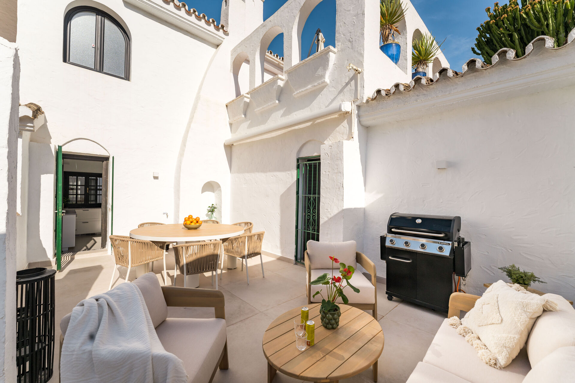 The terrace, with a round dining table, outdoor sofas and a gas barbecue against whitewashed walls, and the kitchen door open on the left.