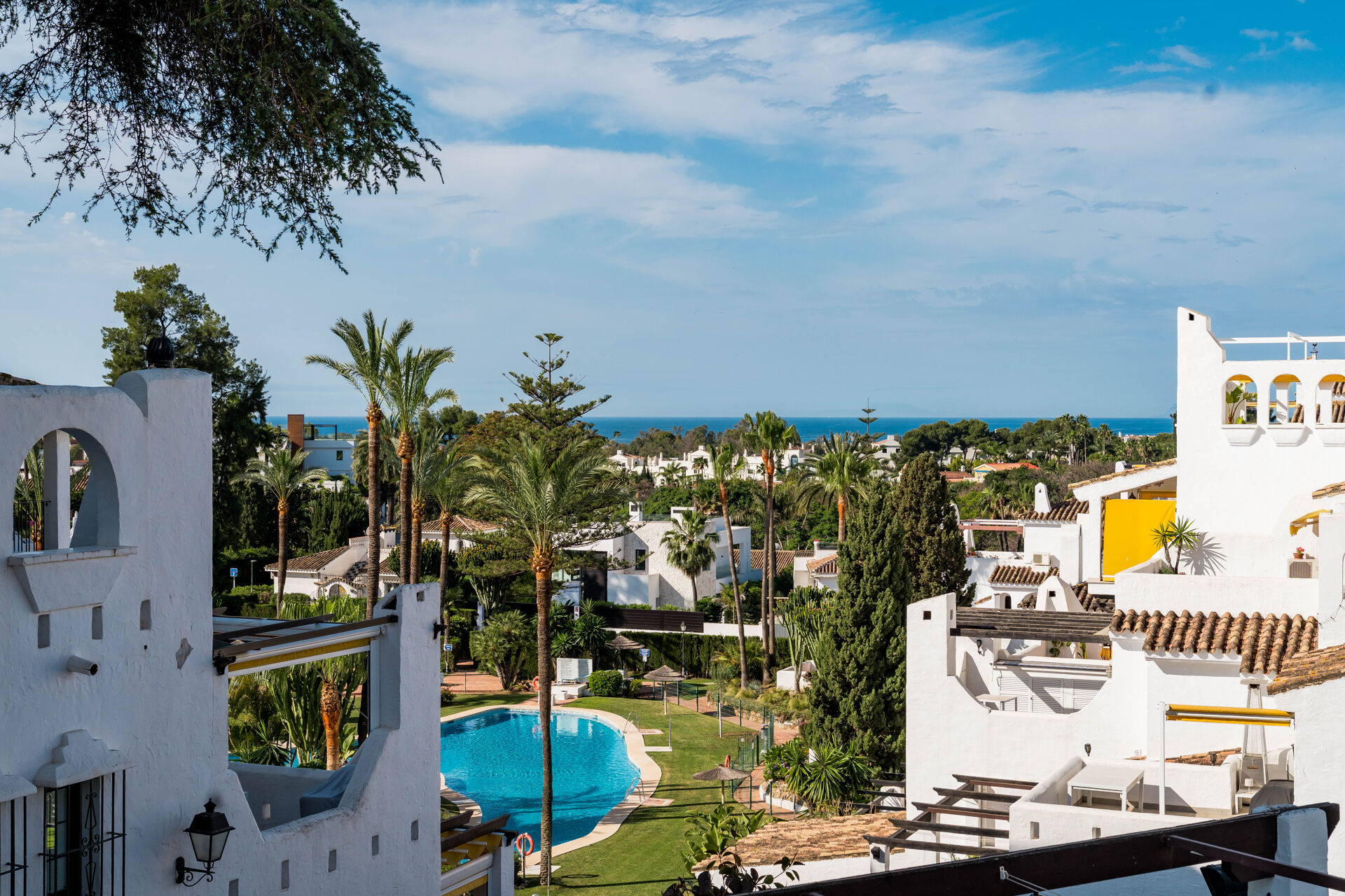 The communal pool and gardens of Aldea Blanca among palm trees and white buildings, with the sea in the distance.