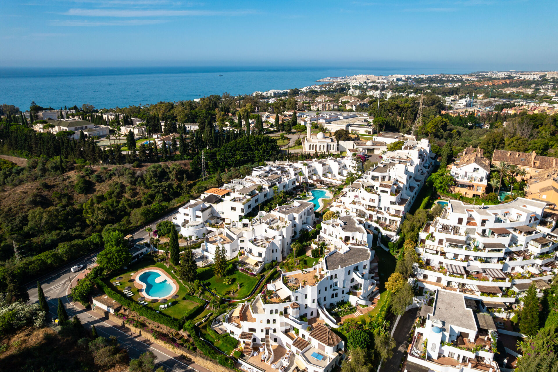 An aerial view of white apartment buildings among gardens and swimming pools, with the coast and the sea beyond.