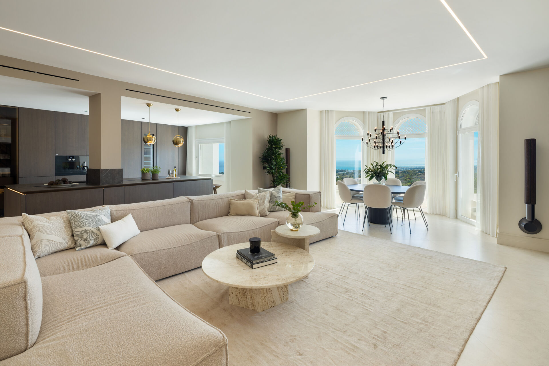 The living room, with a large sofa, the kitchen to the left and the dining table by arched windows looking out to the sea.