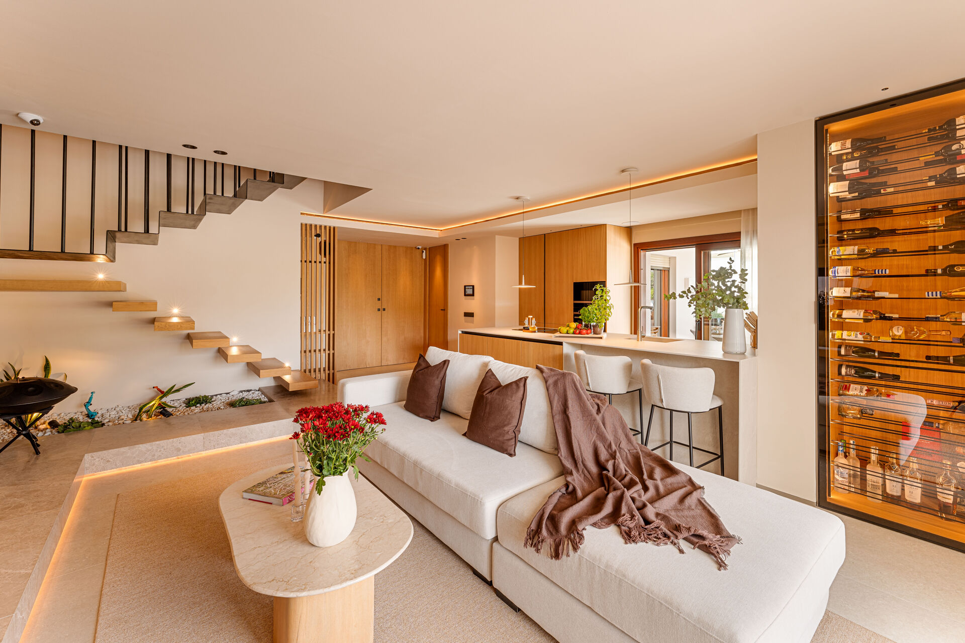 The living room, with a floating timber staircase on the left, the kitchen island behind the sofa and a glass-fronted wine wall on the right.
