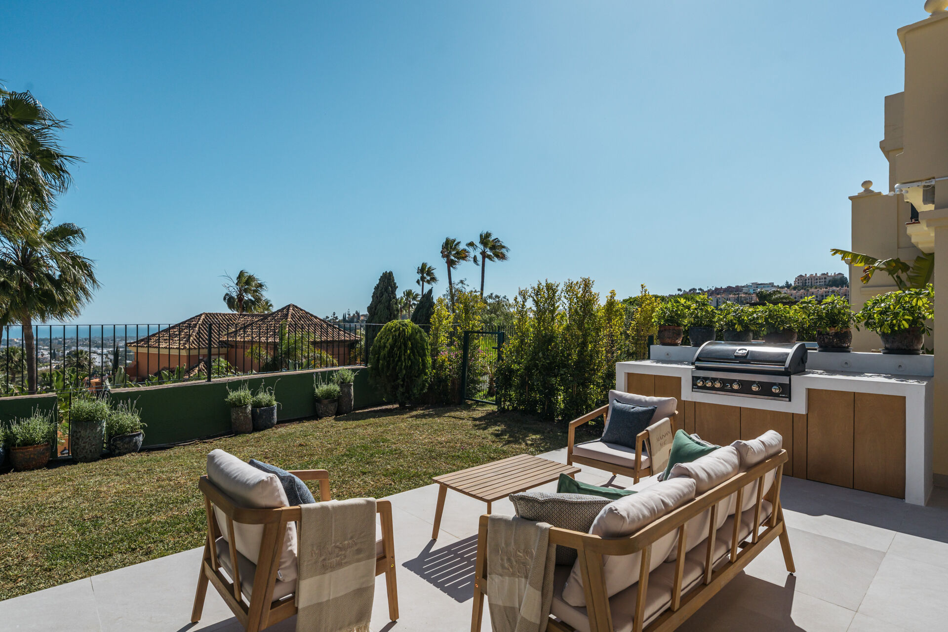 The garden terrace, with an outdoor sofa set and a built-in barbecue beside a lawn edged with hedges and palms.