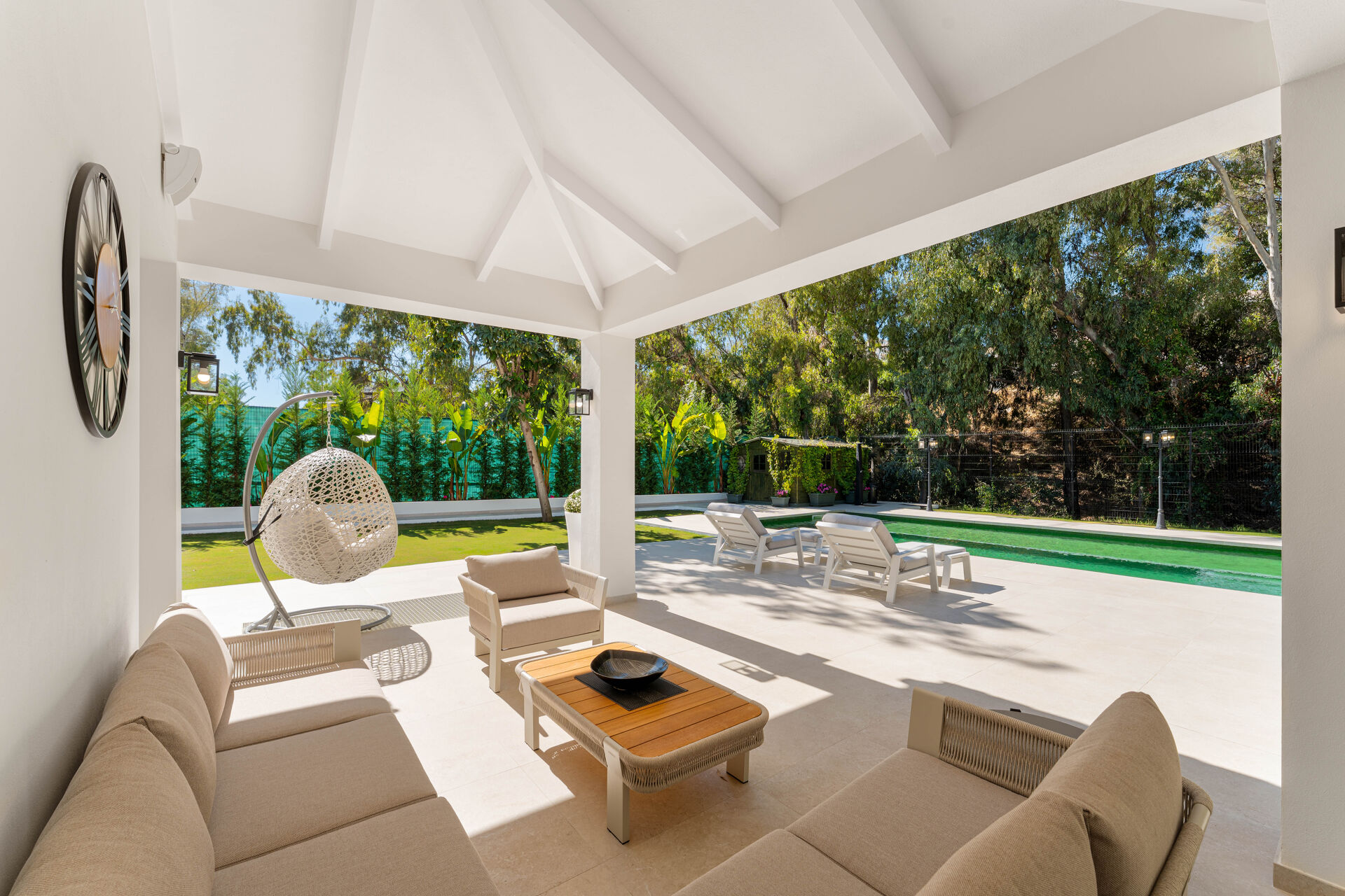The covered terrace with a sofa, an armchair and a hanging chair, looking out to the sun loungers, the pool and the lawn.