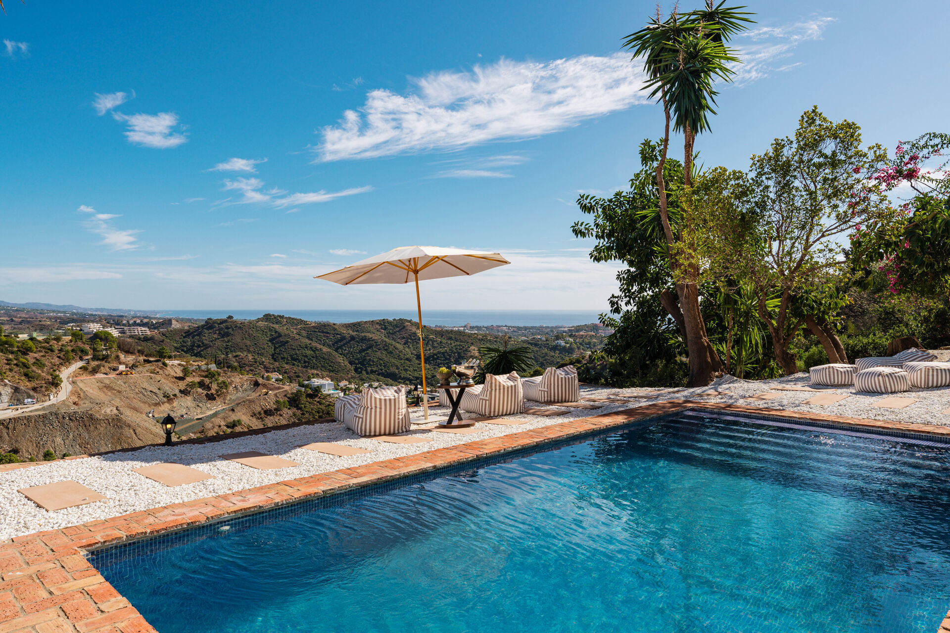 The swimming pool with striped bean bags and a parasol on white gravel, looking over wooded hills to the sea.