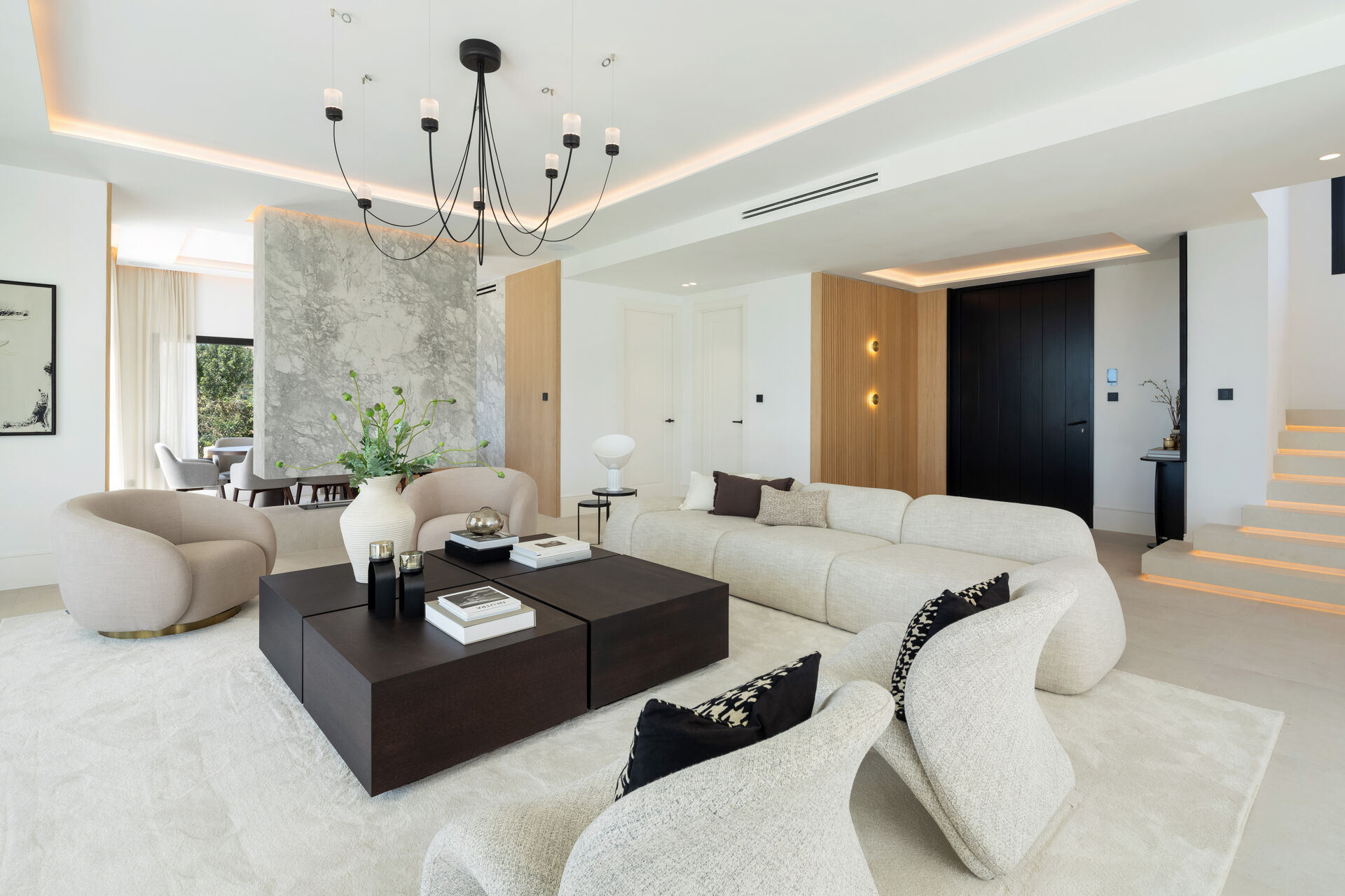 The main living room, with a curved sofa and armchairs, a marble partition and a lit staircase on the right.