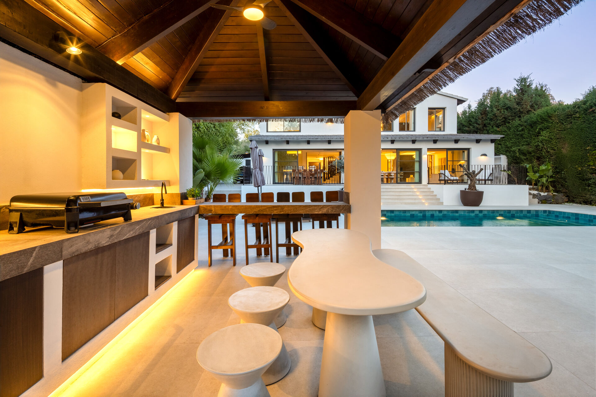The outdoor kitchen under its timber roof at dusk, with a barbecue and a bar, looking across the pool to the back of the villa.