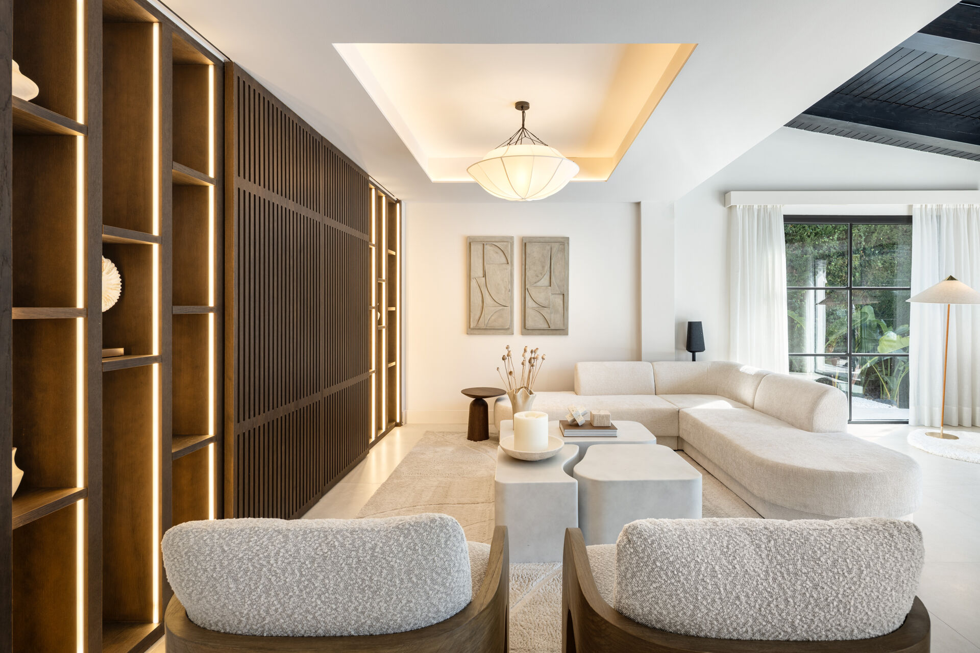 The living room, with a large corner sofa, two armchairs, lit timber shelving and a glass door to the garden.