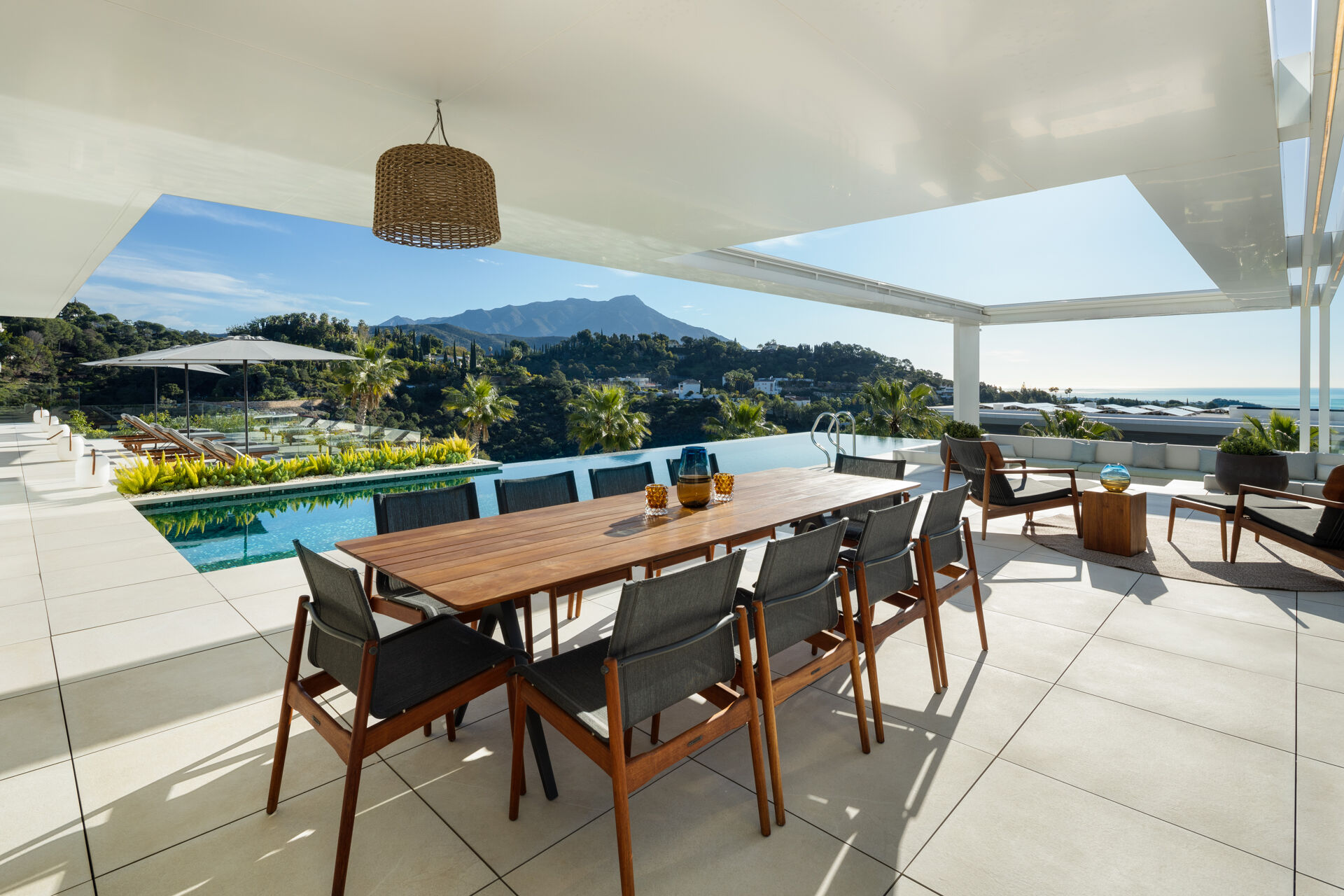 The covered dining terrace beside the infinity pool, with a long timber table and a view over the hills to the mountains and the sea.