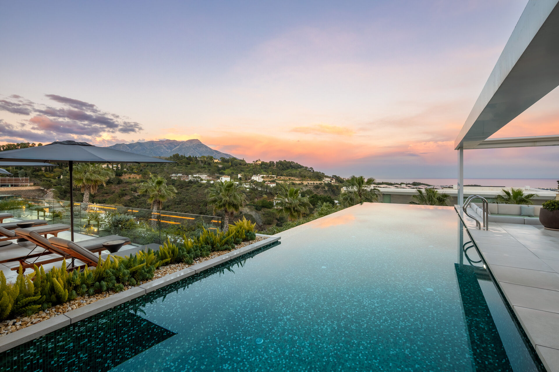 The infinity pool at dusk, with palms and wooded hills below and the mountains and the sea beyond.